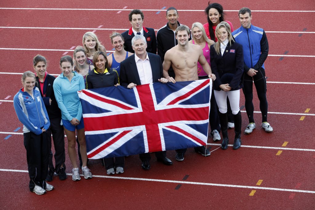 Barrie Wells on a racetrack with a group of young athletes. They are holding up a large Union Jack flag.